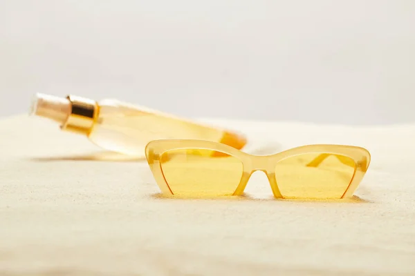 Messa a fuoco selettiva di occhiali da sole gialli vicino bottiglia di olio abbronzante su sabbia isolata su grigio — Foto stock