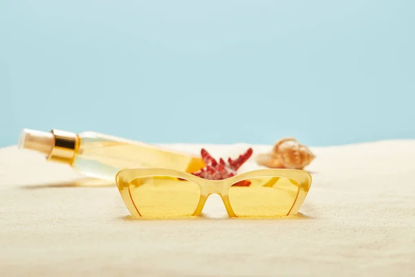 Selective focus of sunglasses near suntan oil bottle, red starfish and seashell on sand  isolated on blue — Stock Photo