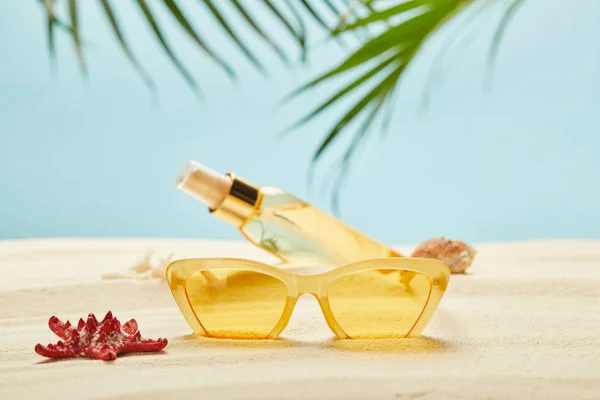 Selective focus of red starfish near sunglasses and bottle with suntan oil on sand isolated on blue — Stock Photo