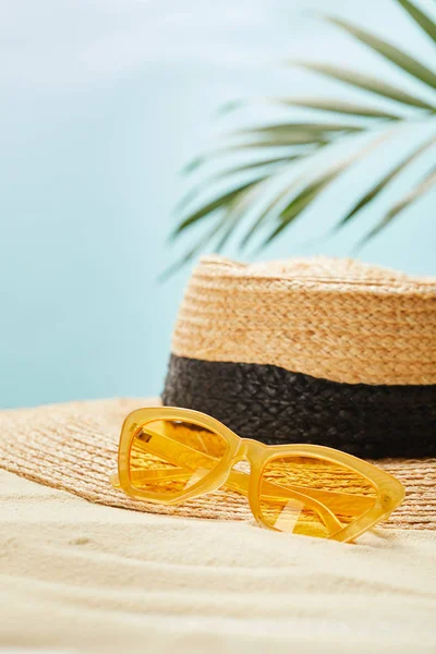 Selective focus of yellow sunglasses near straw hat on golden sand in summertime isolated on blue — Stock Photo
