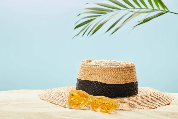 Selective focus of yellow sunglasses near straw hat on sand in summertime isolated on blue — Stock Photo