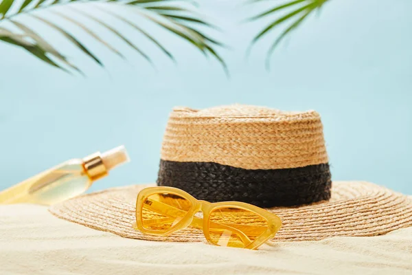 Enfoque selectivo de gafas de sol amarillas cerca de sombrero de paja y botella con aceite bronceador en arena aislada en azul - foto de stock