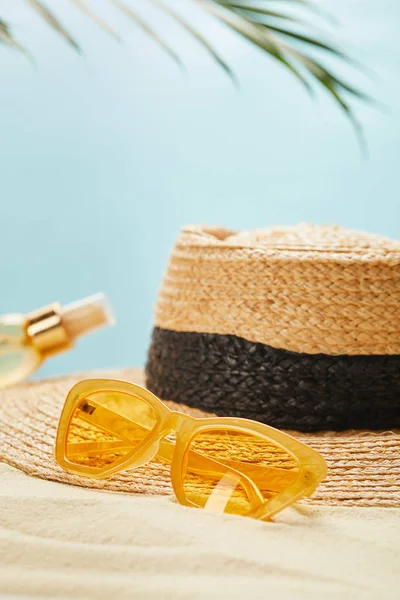 Selective focus of sunglasses near straw hat and bottle with suntan oil on sandy beach isolated on blue — Stock Photo