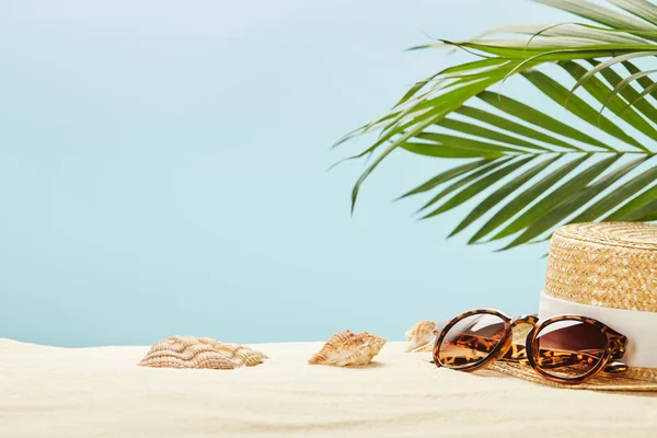 Selective focus of sunglasses and straw hat near seashells and green leaves isolated on blue — Stock Photo
