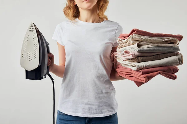 Mulher parcial em t-shirt segurando ferro e roupas passadas dobradas isoladas em cinza — Fotografia de Stock