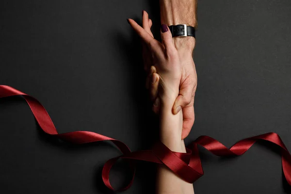 Cropped view of man holding female hand in red satin ribbon on black background — Stock Photo