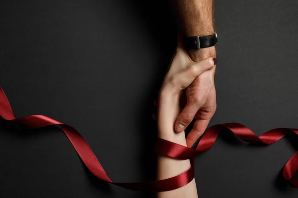 Cropped view of man and woman in red satin ribbon holding hands on black background — Stock Photo