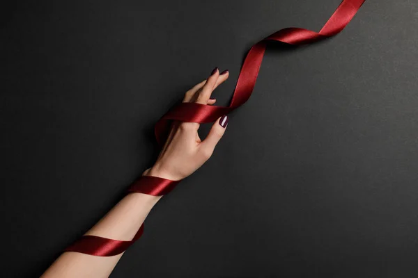 Vista recortada de mano femenina y cinta roja satinada sobre fondo negro - foto de stock