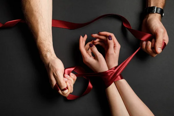 Cropped view of man tying red satin ribbon on female hands on black background — Stock Photo