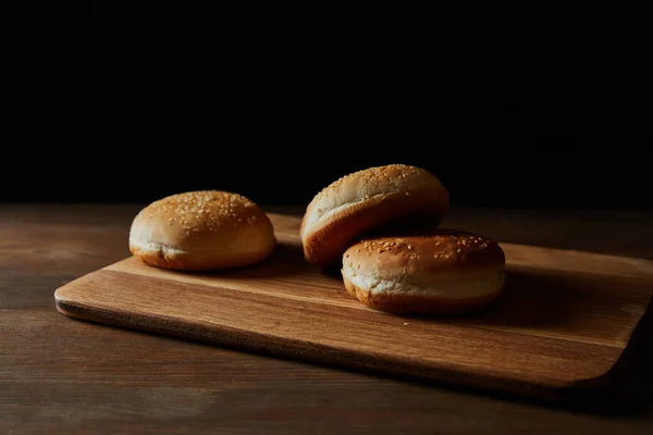 Pães deliciosos frescos com gergelim em tábua de corte de madeira isolada em preto — Fotografia de Stock