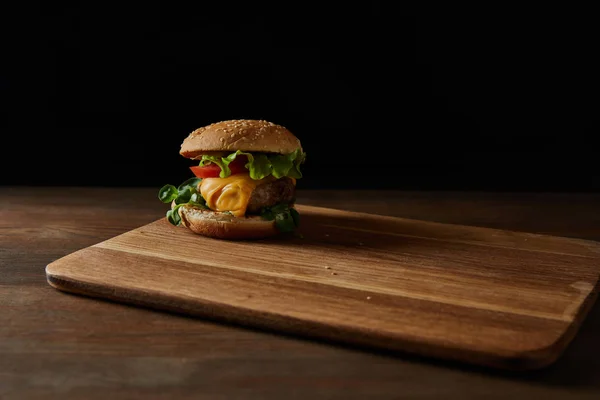 Tasty meat burger with sesame on wooden chopping board isolated on black — Stock Photo