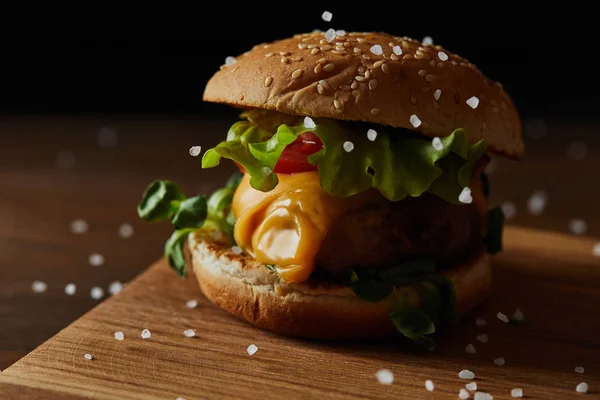 Nahaufnahme von Salz, das auf leckeren Fleisch-Burger mit Käse und Grün auf einem hölzernen Schneidebrett fällt — Stockfoto