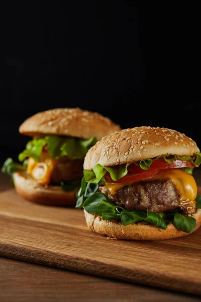 Selective focus of tasty meat burgers on wooden chopping board isolated on black — Stock Photo