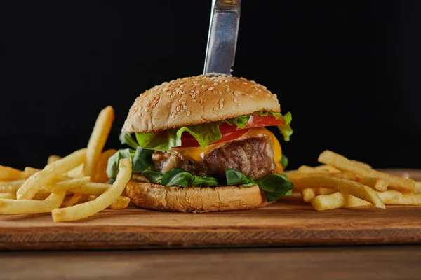 Knife in delicious meat burger with cheese, greenery and sesame near french fries isolated on black — Stock Photo