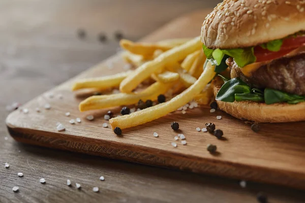 Papas fritas doradas con sabrosa hamburguesa de carne con sal dispersa y pimienta negra sobre tabla de cortar de madera — Stock Photo