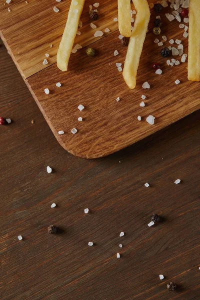 Vue du dessus du sel, du poivre noir et des frites dorées fraîches sur planche à découper en bois — Photo de stock