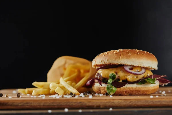 Sale, patatine fritte e delizioso hamburger con carne su superficie di legno isolata su nero — Foto stock