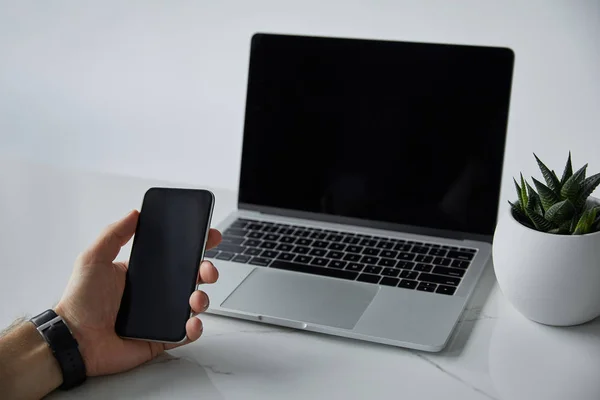 Vista parcial del hombre sosteniendo el teléfono inteligente, portátil con pantalla en blanco y maceta en gris - foto de stock