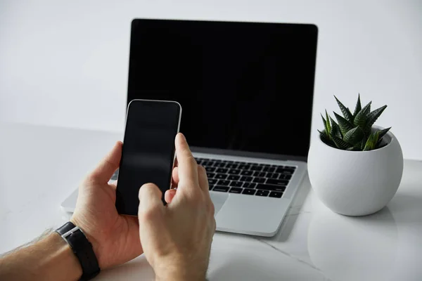 Частковий вид людини, що використовує смартфон біля ноутбука з порожнім екраном, і горщик для квітів ізольований на сірому — стокове фото