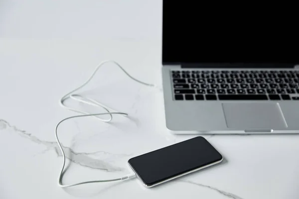 Enfoque selectivo del teléfono inteligente conectado con cable a la computadora portátil con pantalla en blanco aislado en gris — Stock Photo