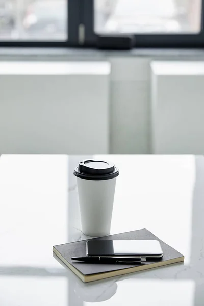 Смартфон с чистым экраном, ноутбуком, ручкой и одноразовой чашкой на белом столе — стоковое фото