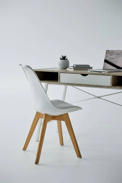Sedia bianca, tavolo da ufficio con laptop, libri e vaso da fiori su grigio — Foto stock