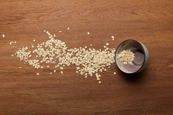 Vista superior de copos de avena esparcidos cerca de cuenco en la mesa de madera - foto de stock