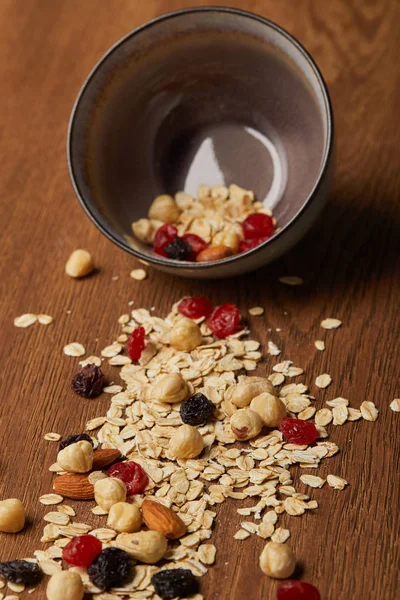 Fiocchi d'avena sparsi con noci e bacche secche dalla ciotola sul tavolo di legno — Foto stock