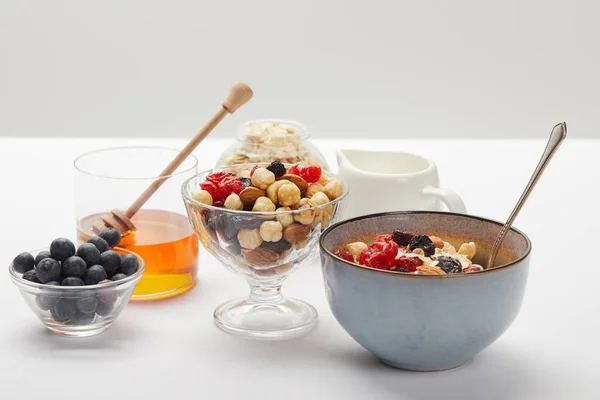 Tazones y vasos con bayas frescas, frutos secos, miel y cereales servidos para el desayuno en mesa blanca aislada en gris - foto de stock