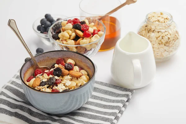 Bowl on striped napkin with oat flakes, nuts and berries on white table with milk jug — Stock Photo