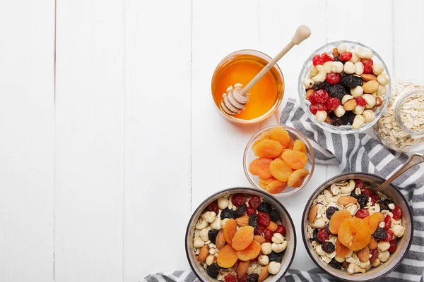 Top view of bowls with muesli, dried berries and nuts served for breakfast with dried apricots and honey on white wooden table — Stock Photo