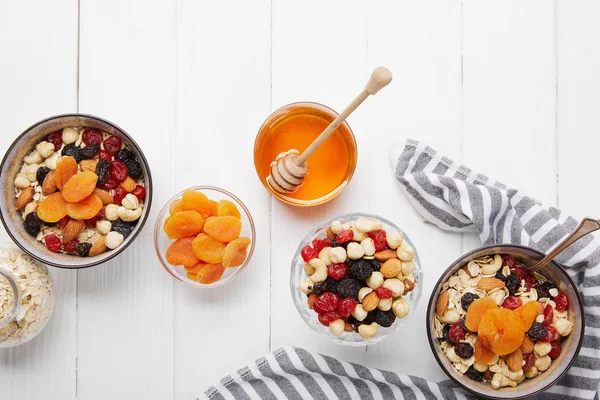 Vista dall'alto di ciotole con cereali, bacche secche e noci servite per colazione con albicocche secche e miele vicino a un panno rigato su un tavolo di legno bianco — Foto stock