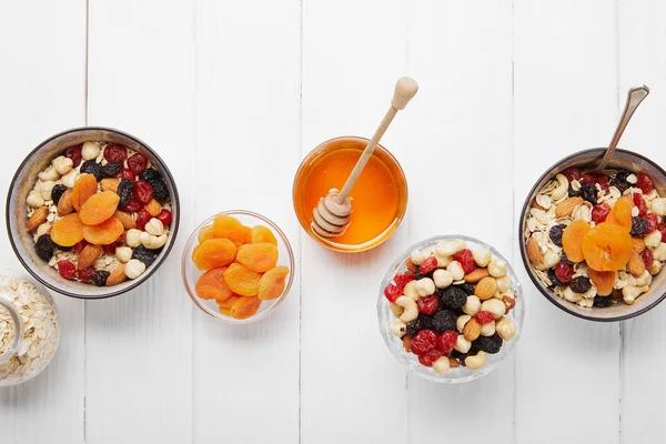 Top view of bowls with cereal, dried apricots and berries, honey and nuts served for breakfast on white wooden table — Stock Photo