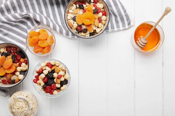 Top view of bowls with cereal, dried apricots and berries, honey and nuts on white table with striped napkin — Stock Photo