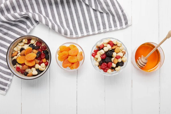 Top view of bowls with cereal, dried apricots and berries, honey and nuts on white table with striped cloth — Stock Photo