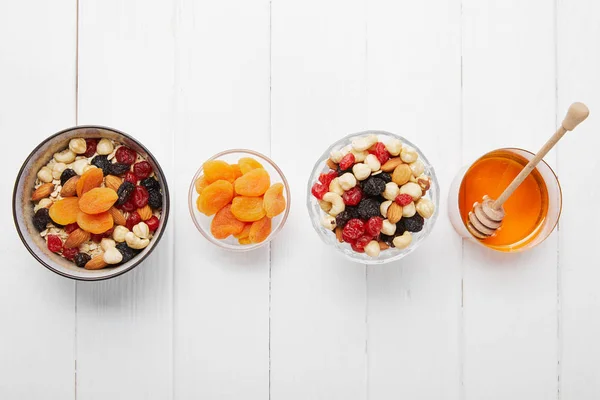 Top view of bowls with cereal, dried apricots and berries, honey and nuts in row on white table — Stock Photo