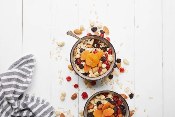 Vista dall'alto di ciotole con muesli, albicocche secche e bacche e noci servite a colazione con ingredienti sparsi sul tavolo di legno bianco — Foto stock