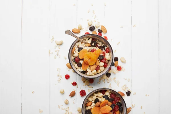 Top view of bowls with muesli, dried apricots and berries and nuts served for breakfast with scattered ingredients on white table — Stock Photo