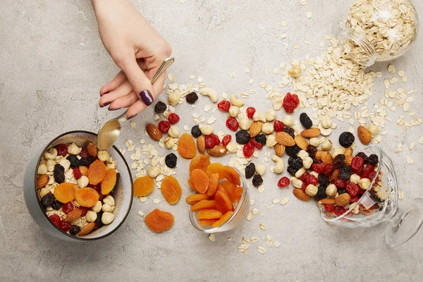 Cropped view of woman holding spoon near bowls with muesli, dried apricots and berries, nuts on textured grey surface with messy scattered ingredients — Stock Photo
