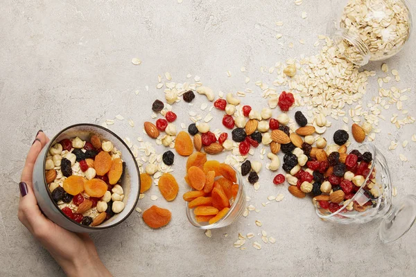 Vista recortada de la mujer sosteniendo tazón con muesli, albaricoques secos y bayas, nueces en la superficie gris texturizada con ingredientes dispersos desordenados - foto de stock