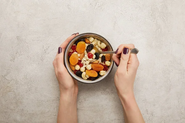 Vista recortada de la mujer sosteniendo cuchara y tazón con muesli, albaricoques secos y bayas, nueces en la superficie gris texturizada - foto de stock