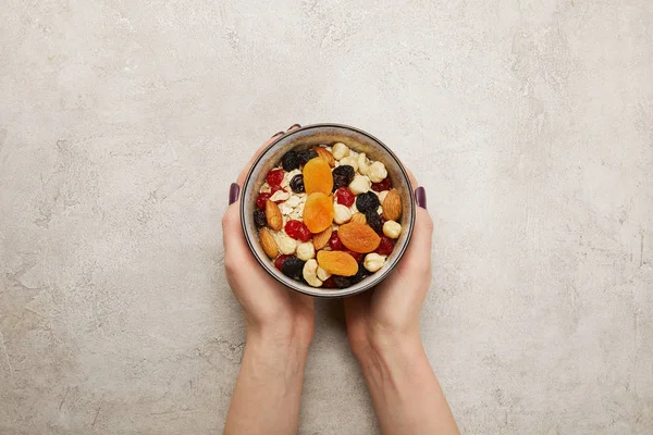 Vista recortada de la mujer sosteniendo tazón con muesli, albaricoques secos y bayas, frutos secos en la superficie gris texturizada - foto de stock