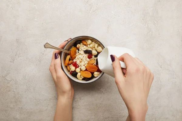 Vista ritagliata della donna versando il latte dalla brocca di latte in ciotola con muesli, albicocche secche e bacche, noci su superficie grigia testurizzata — Foto stock