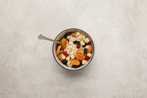 Vista dall'alto della ciotola con muesli, albicocche secche e bacche, noci e cucchiaio su superficie grigia testurizzata — Stock Photo