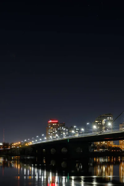 Dunkles Stadtbild mit beleuchteten Gebäuden, Lichtern, Brücke und Fluss — Stockfoto