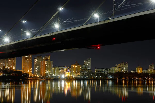 Темний міський пейзаж з мостом, відображення на річці та освітлені будинки вночі — стокове фото
