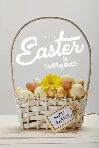 Cesta de mimbre con coloridos huevos pintados, flor de gerberas amarillas, tarjeta de felicitación y feliz Pascua a todo el mundo con letras sobre fondo gris - foto de stock