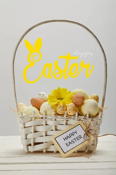 Panier en osier avec œufs peints colorés, fleur de gerbera jaune, carte de vœux et joyeux lettrage de Pâques sur fond gris — Photo de stock