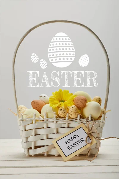 Cesta de mimbre con huevos de colores, flor de gerberas amarillas, tarjeta de felicitación y letras de Pascua sobre fondo gris - foto de stock