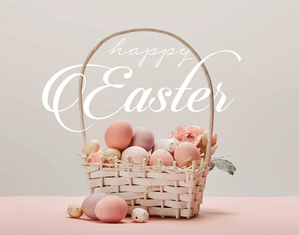 Wicker basket with pink painted eggs, flowers and happy Easter white lettering on grey background — Stock Photo